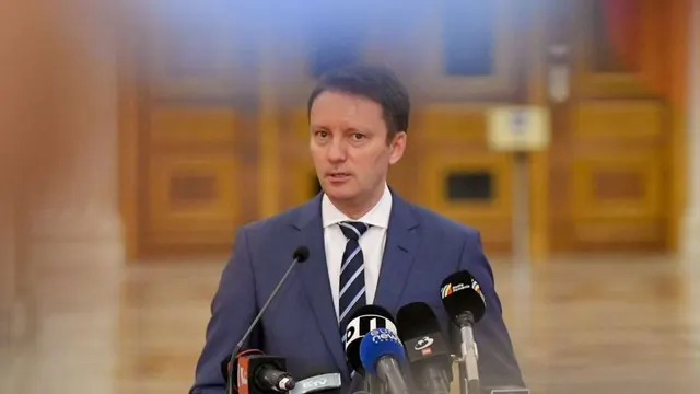 Bărbat tânăr în costum, cu cravată în dungi, vorbind la mai multe microfoane, în fața unor uși de lemn