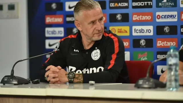 Bărbat cu părul grizonat, în trening negru, la o conferință de presă în fața panoului FCSB