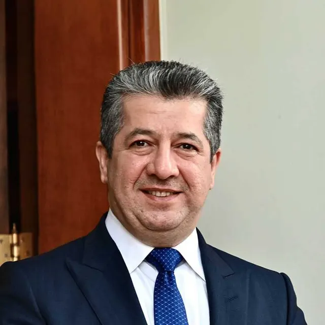 Bărbat zâmbitor, cu părul grizonat, în costum și cravată albastră, lângă o ușă de lemn