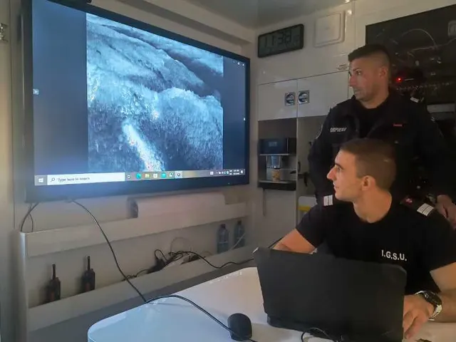 Doi angajați IGSU urmărind pe un ecran mare imagini aeriene ale unui versant, într-un centru de comandă