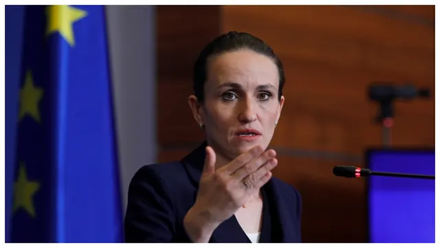 Femeie cu părul prins, în sacou închis, gesticulând lângă drapelul UE și un microfon