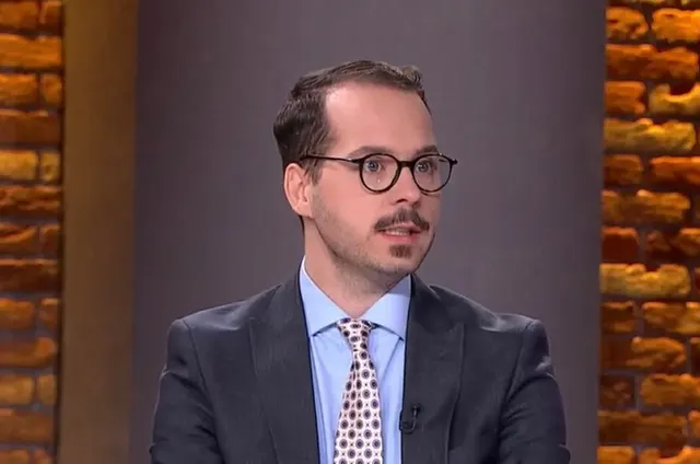 Bărbat cu ochelari rotunzi și mustață, în costum, într-un studio TV cu perete de cărămidă