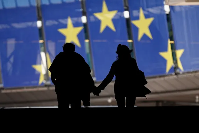 Siluetele a doi oameni ținându-se de mână, în fața unor drapele mari ale UE