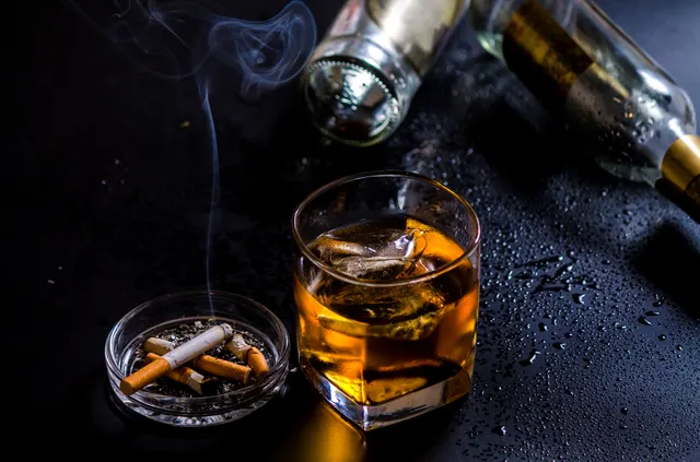 Pahar de whisky cu gheață lângă o scrumieră cu țigări și sticle, pe o masă întunecată