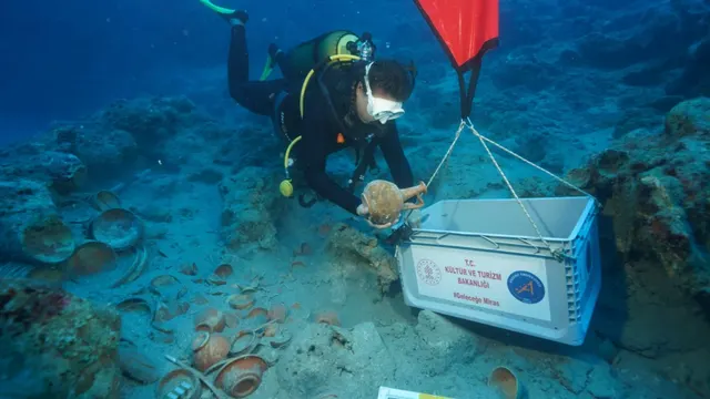 Scafandru care așază un vas antic de ceramică într-o ladă, lângă alte vase de pe fundul mării