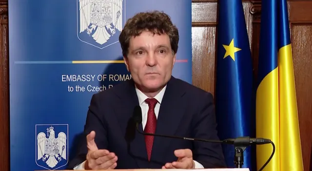 Bărbat în costum vorbind la un microfon, în fața panoului Ambasadei României în Cehia și a drapelelor