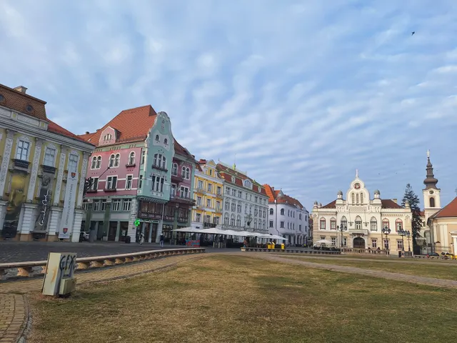 Piață pavată cu clădiri istorice colorate și un turn de biserică, sub un cer cu nori