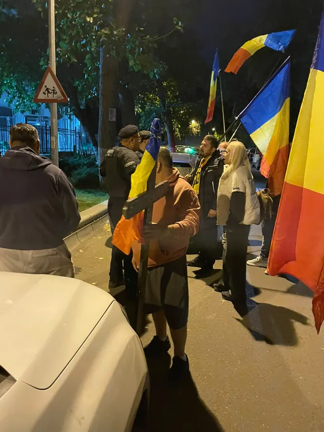 Oameni cu steaguri tricolore adunați noaptea pe un trotuar, lângă mașini parcate