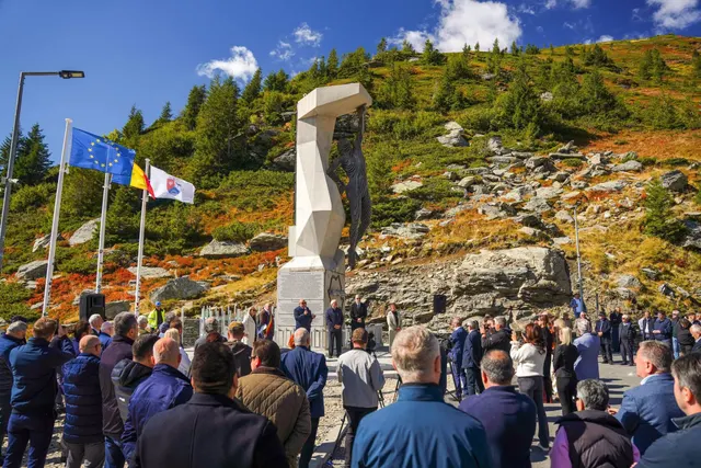 Monumentul alb de pe Transfăgărășan, pe un versant, cu o mulțime adunată și drapelele României și UE