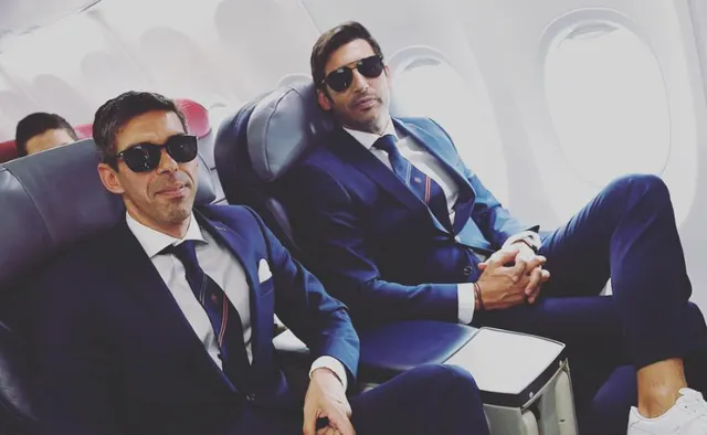 Doi bărbați în costume și ochelari de soare, așezați pe scaune de avion