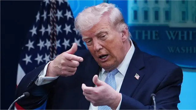 Bărbat cu părul blond-cărunt gesticulând cu ambele mâini, în fața drapelului SUA și a unui panou „The White House”