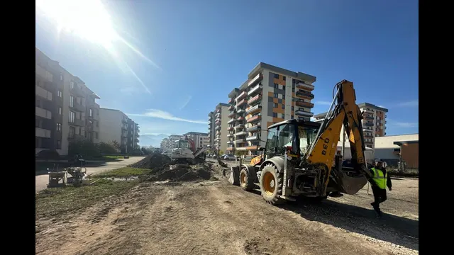 Buldoexcavator galben lucrând pe un teren de pământ, între blocuri noi, cu un muncitor în vestă