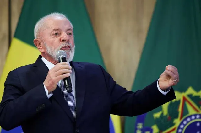 Bărbat cu barbă albă, în costum, vorbind la microfon și gesticulând, în fața drapelului Braziliei