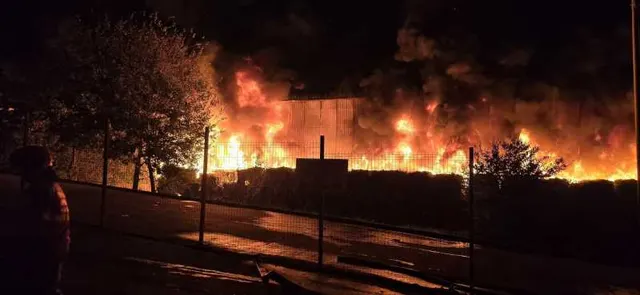 Flăcări uriașe noaptea deasupra unei hale, văzute din spatele unui gard