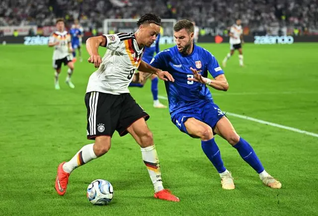 Fotbalist în tricou alb și șort negru driblând un adversar în echipament albastru, pe un stadion plin