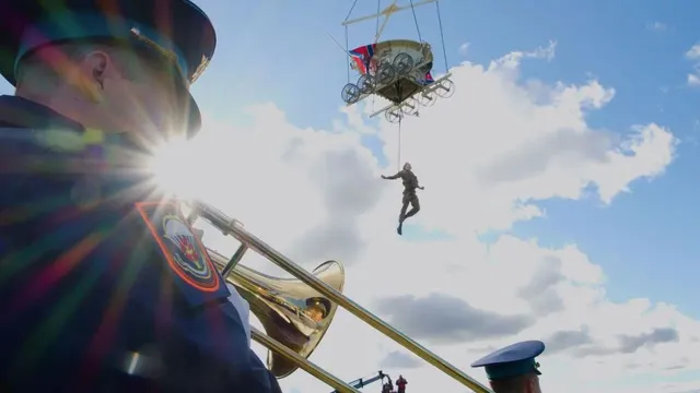 Muzician cu trombon în prim-plan; pe cer, un balon ridică o platformă cu o siluetă agățată dedesubt