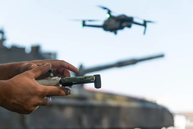Mâini care țin telecomanda unei drone aflate în zbor, cu țeava unui tanc în fundal