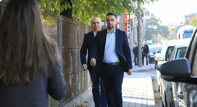 Doi bărbați în costume mergând pe un trotuar lângă un gard, cu o femeie cu spatele în prim-plan