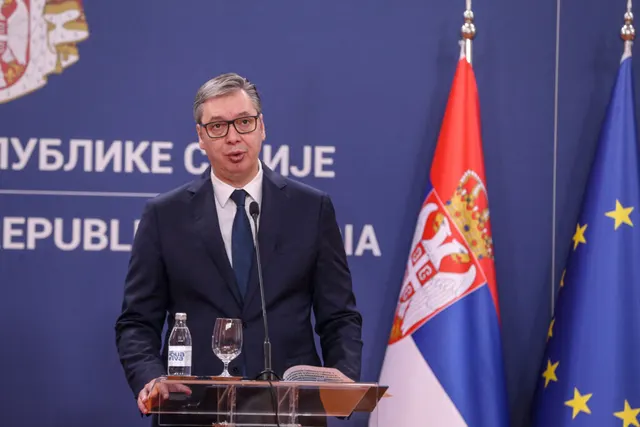 Bărbat în costum la un pupitru, între drapelele Serbiei și UE, în fața unui panou cu stema Serbiei
