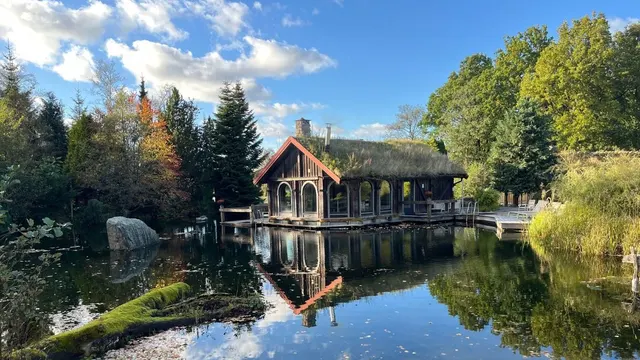 Casă de lemn pe malul unui lac, între brazi și copaci cu frunze de toamnă, oglindită în apă