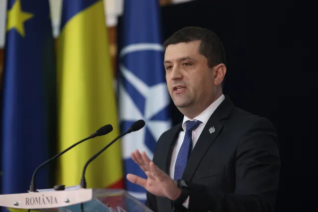 Bărbat în costum vorbind la un pupitru cu inscripția „România”, în fața drapelelor UE, României și NATO