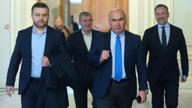 Patru bărbați în costume mergând pe un hol; în centru, unul cu cravată albastră