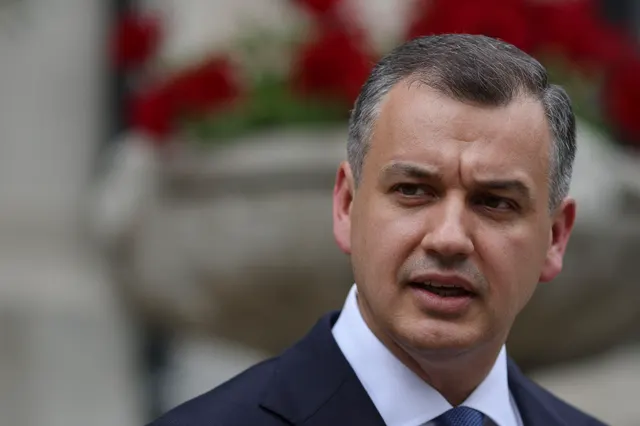 Bărbat în costum, cu părul grizonat, vorbind, cu flori roșii în fundal