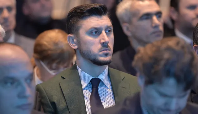 Bărbat cu barbă, în costum și cravată, așezat în public printre alți participanți