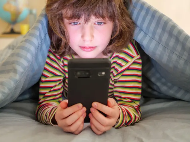 Copil sub o pătură, cu fața luminată de ecranul unui telefon