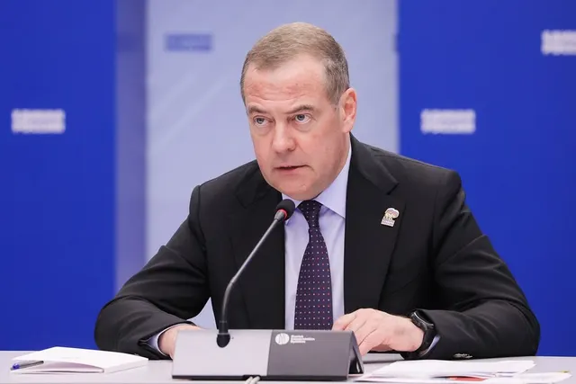 Bărbat în costum închis și cravată, vorbind la un microfon de la o masă, pe fundal albastru