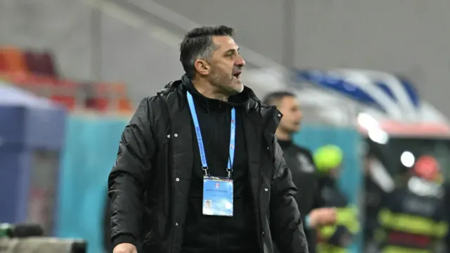 Bărbat cu barbă, în geacă neagră și cu acreditare la gât, pe marginea unui teren