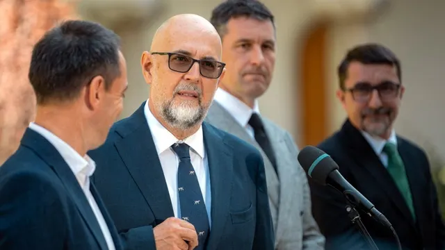 Patru bărbați în costume în fața microfoanelor; în centru, unul chel, cu barbă și ochelari fumurii