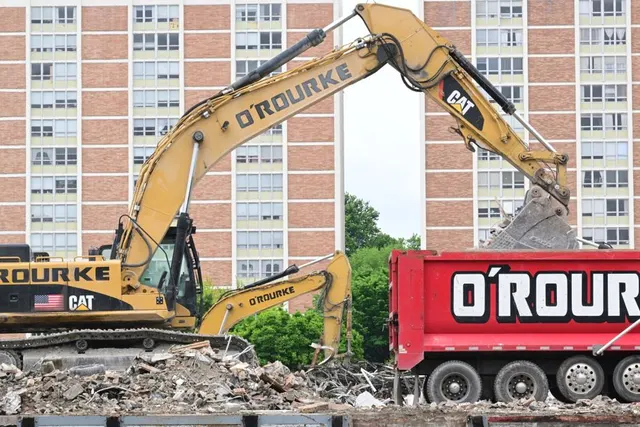 Excavator galben care demolează o clădire, lângă un camion roșu O'Rourke și blocuri înalte de locuințe