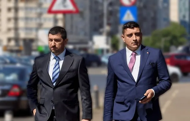 Doi bărbați în costume închise la culoare, mergând pe un trotuar, cu mașini parcate în spate