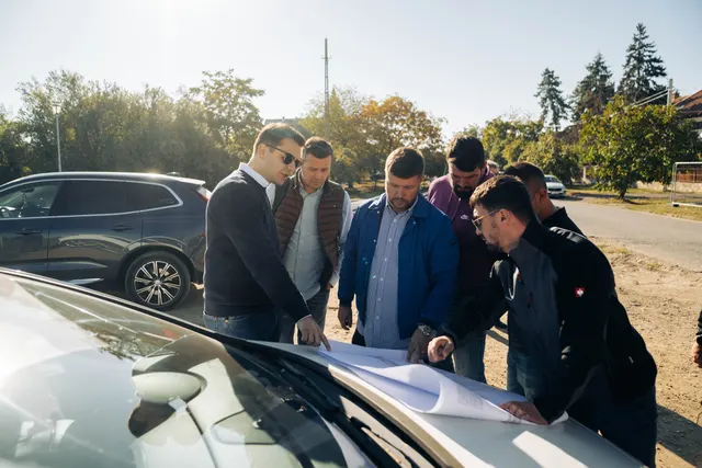 Bărbați în costume privind planuri întinse pe capota unei mașini, la începutul unui șantier