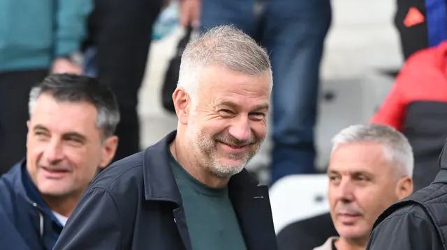 Bărbat cu părul grizonat și barbă, zâmbind, între doi bărbați, într-o tribună de stadion