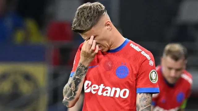 Fotbalist în tricoul roșu al FCSB, cu mâna la ochi, pe teren, cu un coechipier în spate