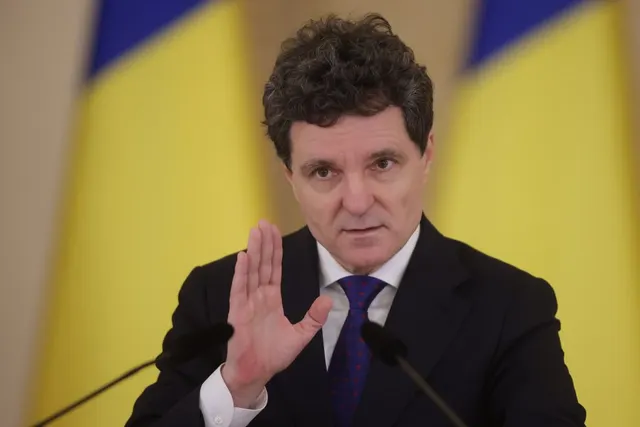 Bărbat cu părul creț, în costum, cu palma ridicată, vorbind la microfon între drapelele României