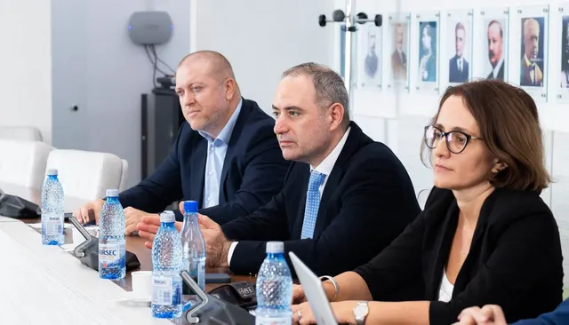 Trei oficiali, doi bărbați și o femeie cu ochelari, la o masă de ședință, cu portrete pe perete în spate