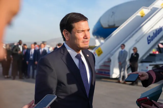 Bărbat în costum închis lângă scara unui avion, înconjurat de oameni cu telefoane, pe o pistă