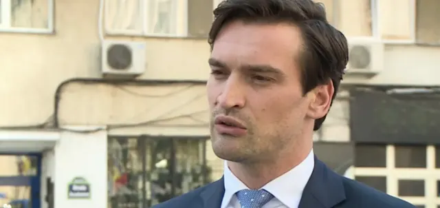 Bărbat brunet, în costum și cămașă albă, vorbind într-un interviu pe o stradă