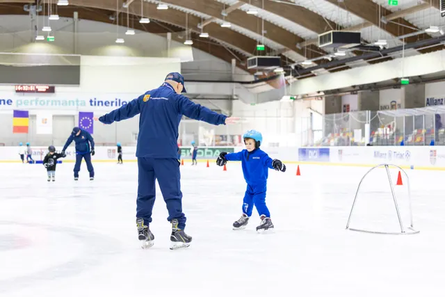Instructor și un copil cu cască patinând pe un patinoar acoperit, lângă o poartă de hochei