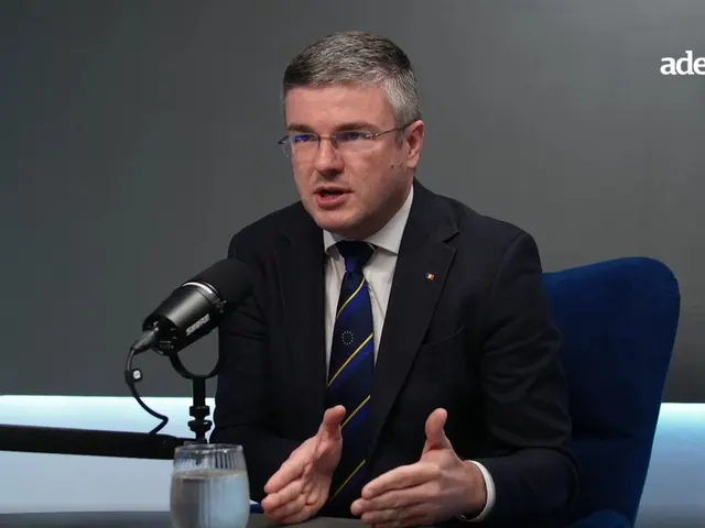 Bărbat cu ochelari, în costum, vorbind la un microfon de studio, cu mâinile deschise