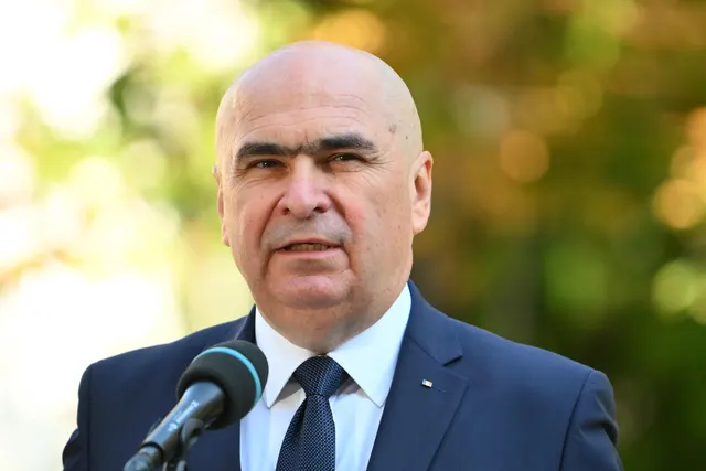 Bărbat chel, în costum bleumarin și cravată, vorbind la un microfon, pe fundal de copaci înverziți