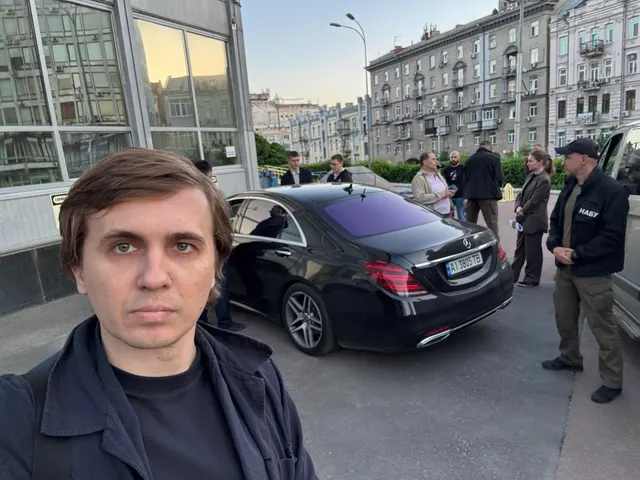 Mihailo Tkaci, în geacă neagră, pe o stradă, cu o mașină neagră și oameni în spate