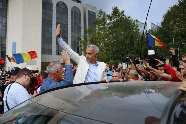 Călin Georgescu, în sacou alb, salutând cu mâna ridicată o mulțime cu drapele tricolore