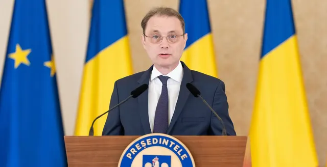 Bărbat cu ochelari vorbind la pupitrul Președinției României, între drapelele UE și României