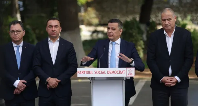 Bărbat vorbind la un pupitru al Partidului Social Democrat, în aer liber, cu alți trei bărbați lângă el