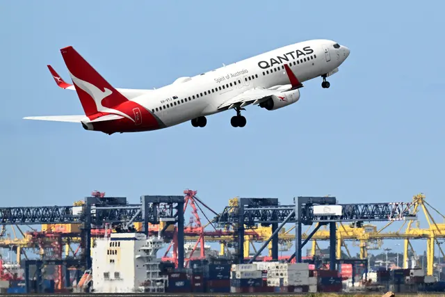 Avion Qantas, cu coada roșie și cangurul alb, decolând deasupra unui port de containere