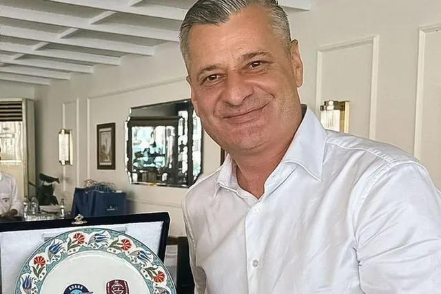 Bărbat cu părul grizonat, în cămașă albă, zâmbind într-un birou, lângă o farfurie decorativă cu o emblemă